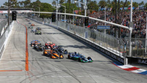 Formula race cars speeding off from a street circuit start, with a full grandstand to the right and palm trees in the background on Aquarium Way