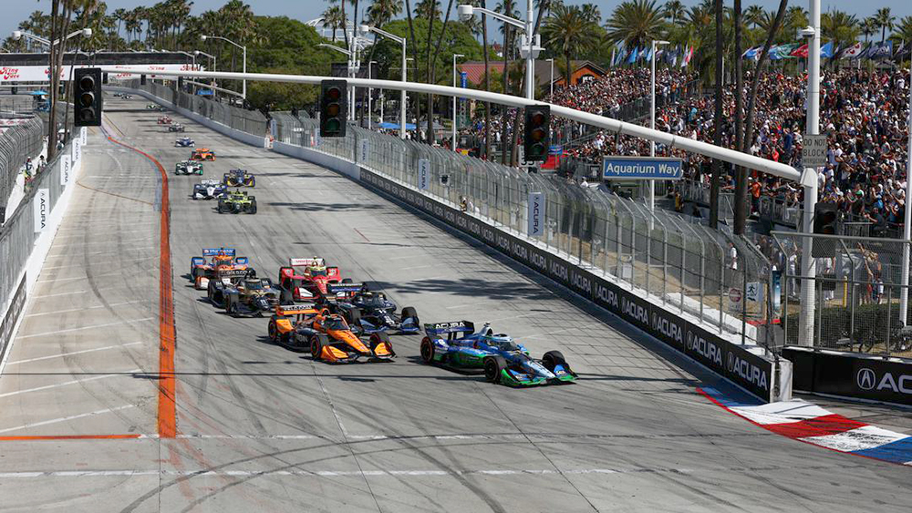 Formula race cars speeding off from a street circuit start, with a full grandstand to the right and palm trees in the background on Aquarium Way