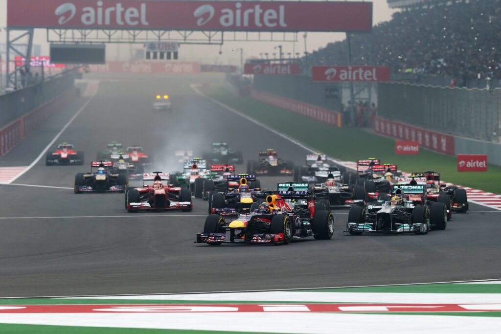 Multiple Formula 1 cars race off the start line on a track with sponsor banners above and a crowded grandstand in the background on a hazy day.
